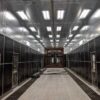 Inside a Kresco H-style blast room: rubber-lined walls, LED lighting, overhead monorail with suspended weldment, and full-floor media recovery grating