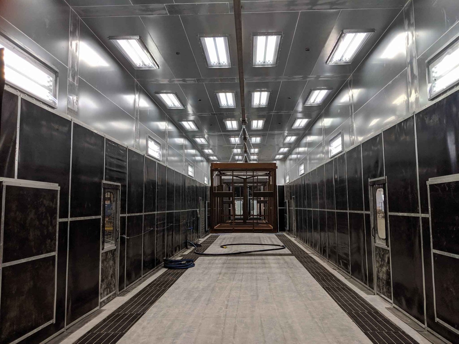 Inside a Kresco H-style blast room: rubber-lined walls, LED lighting, overhead monorail with suspended weldment, and full-floor media recovery grating