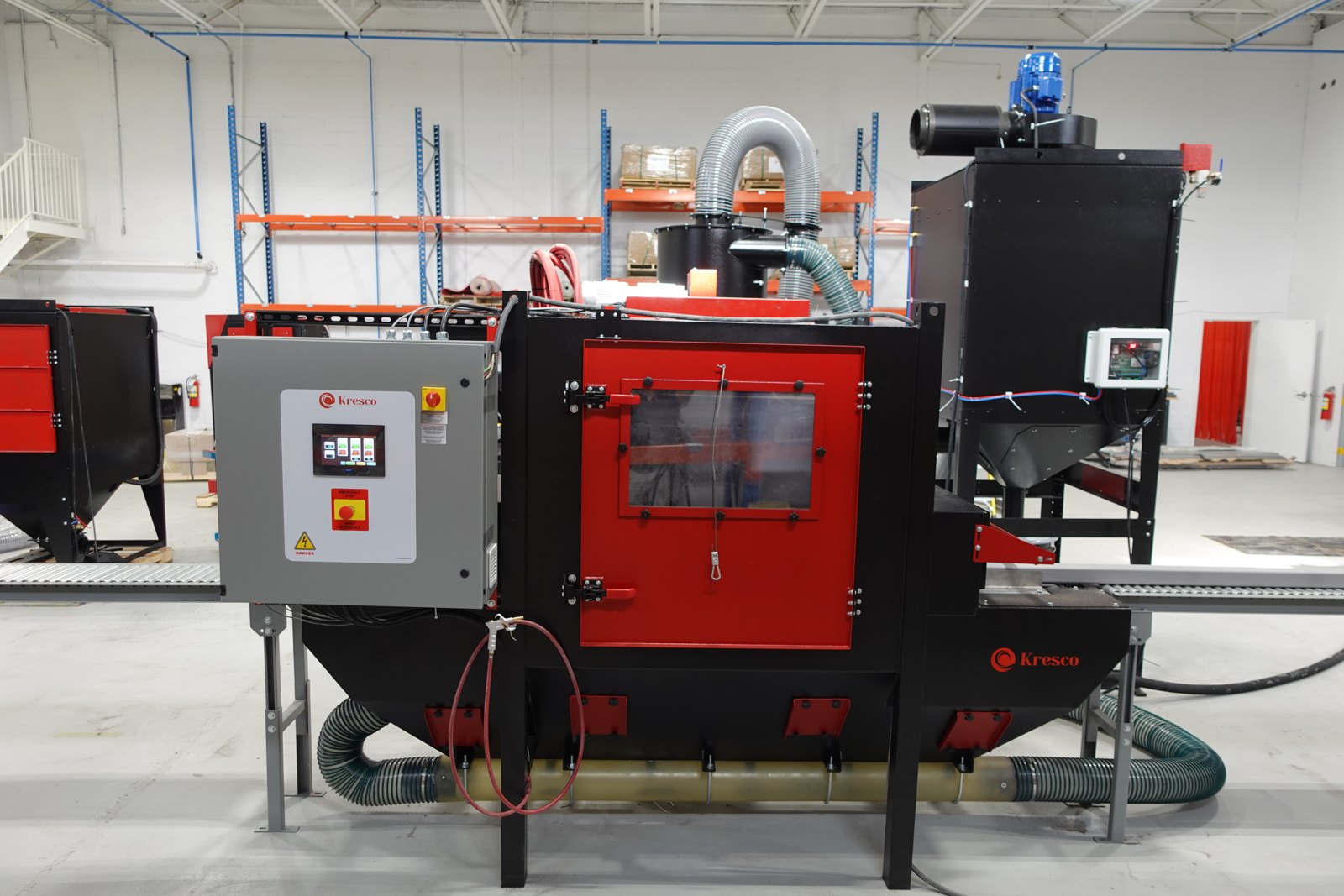 Kresco inline conveyor blast cabinet, front view: roller conveyor pass-through, touchscreen PLC control panel with emergency stop, cyclone media reclaim and cartridge dust collector